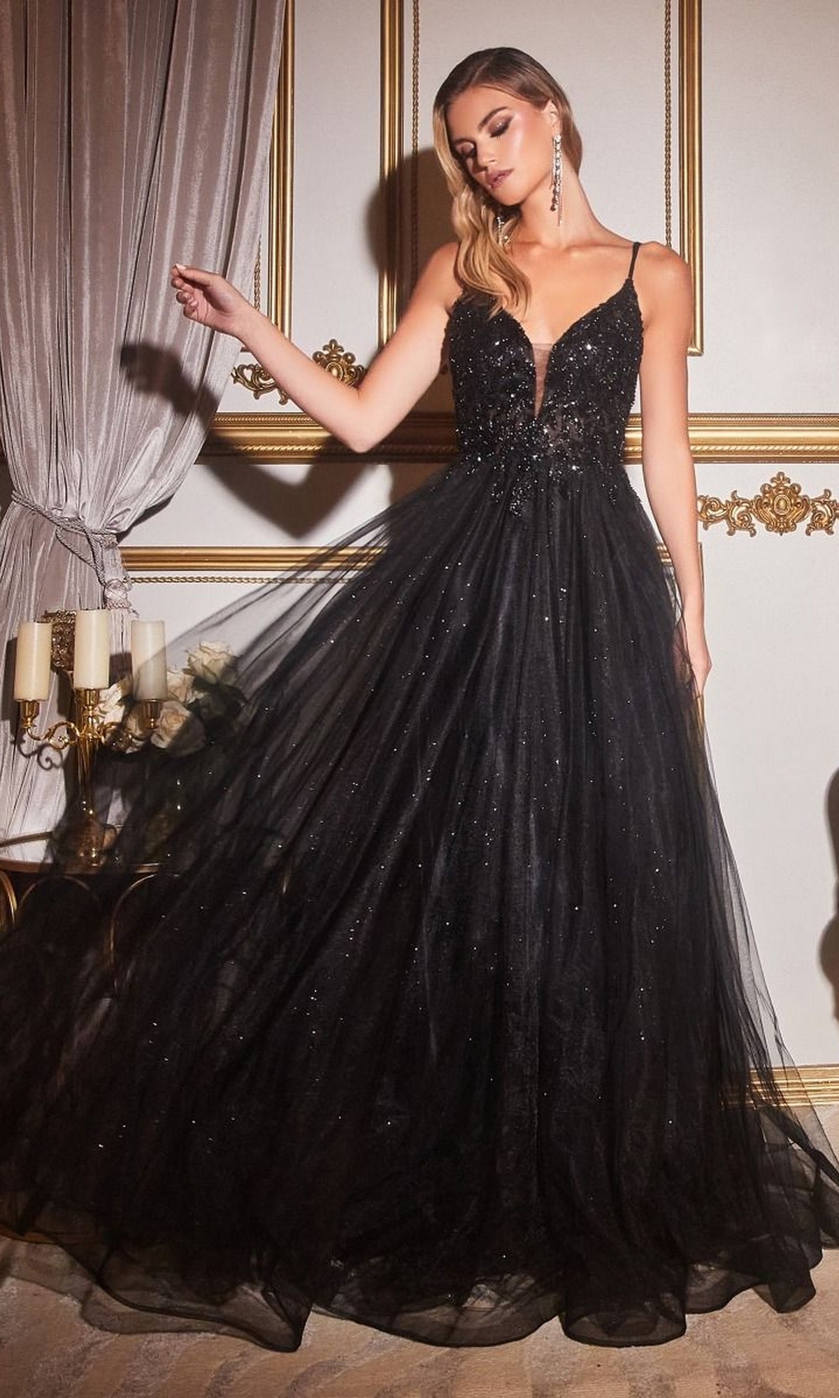 A woman in the Sheer-Corset Long Prom Ball Gown CD0154 in black, featuring a beaded plunging bodice, thin straps, and a full glitter-tulle skirt, poses in an ornate room with gold-trimmed walls.